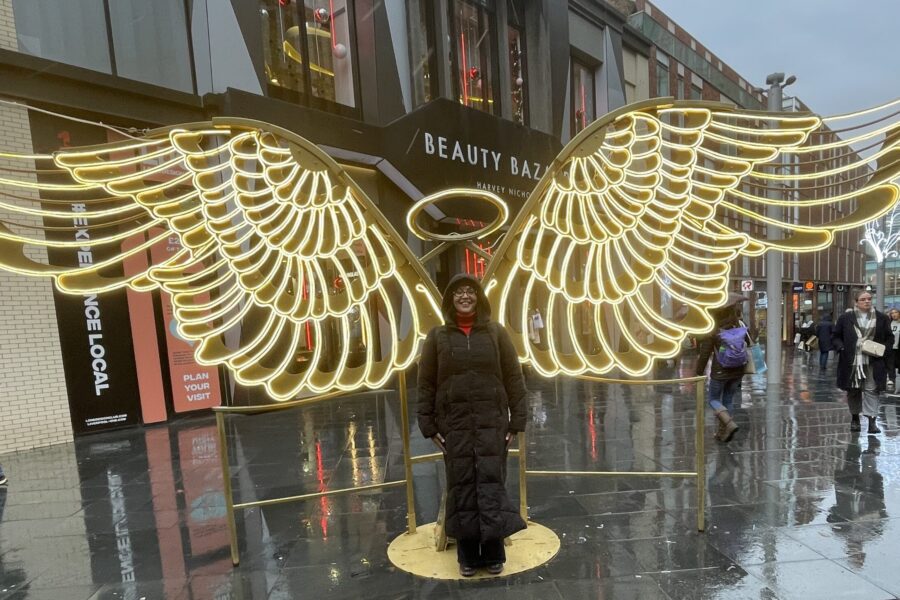 Sharon Hogson standing in the middle of a pedenstrianised street with shops ether side, with illuminated angel wings and a halo behind her.