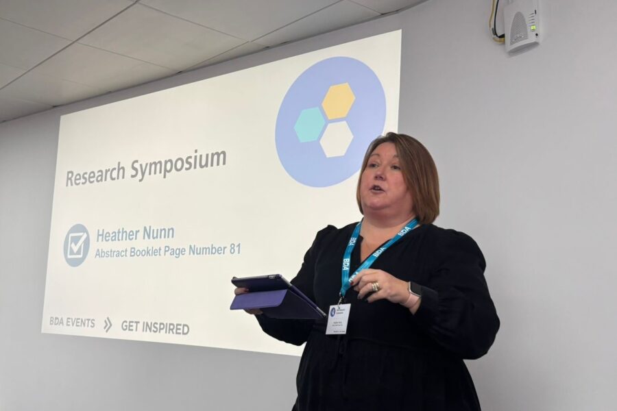 Heather Nunn, standing in front of a white wall with her presentation projected on to it, holding a tablet in her right hand as she talks.