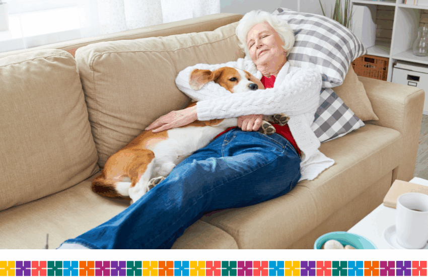 An elderly lady laying on a light brown sofa while cuddling her brown and white dog.