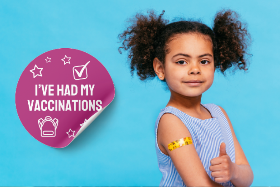 Young girl looking at the camera with her thumb up. She has a yellow sticker on her arm. Next to her is a big purple sticker that says