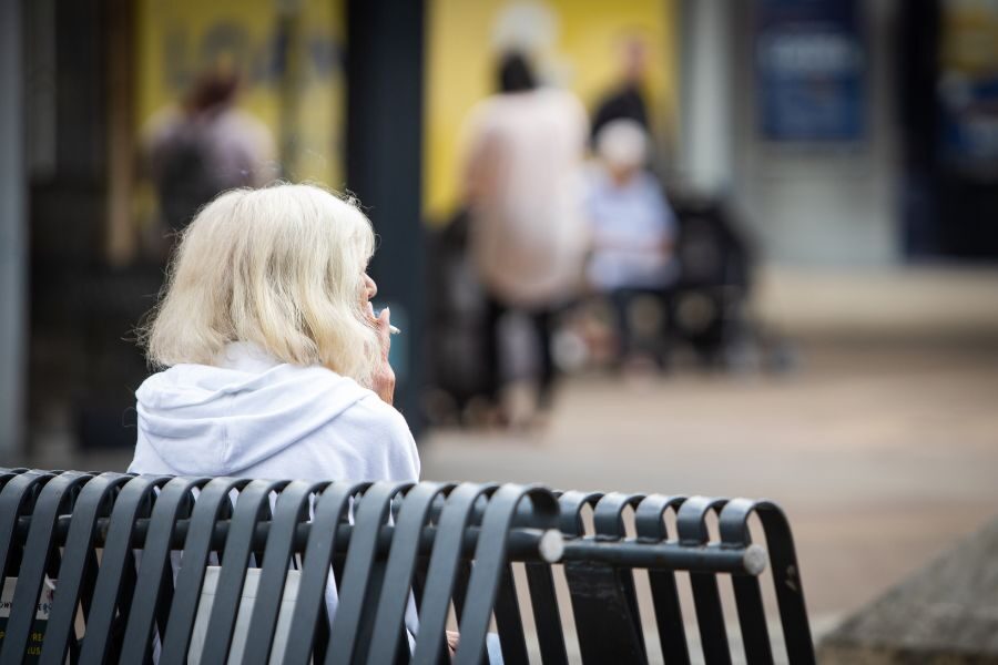 Older lady sat on bench outside smoking a cigarette
